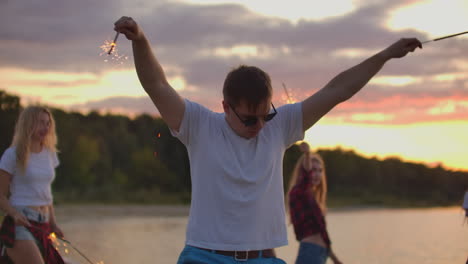 The-brutal-man-with-black-sun-glasses-is-dancing-with-big-bengal-lights-on-the-sand-beach-with-his-friends.-This-is-wonderful-summer-evening-on-the-open-air-party.