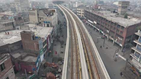 Aerial-Flying-Over-Along-Empty-Orange-Metro-Railtrack-In-Lahore-Towards-Elevated-Station-And-Over-Rooftop