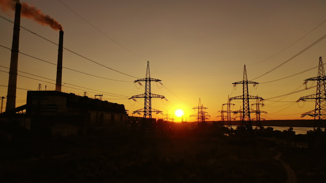 Industrial zone at sunset. High-voltage electric lines near factory with pipes with dirty smoke in the evening. Dark silhouettes of power station.