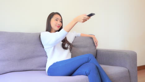 pretty asian woman sitting in sofa, changing television channels with tv remote, full frame slow motion