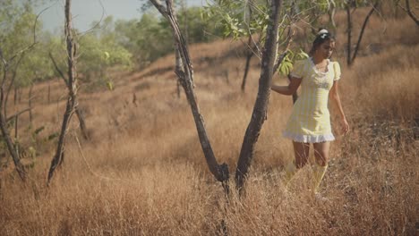 serene footage of a young beautiful indian girl walking coyly around a tree on a hill in a field of tall dead yellow grass