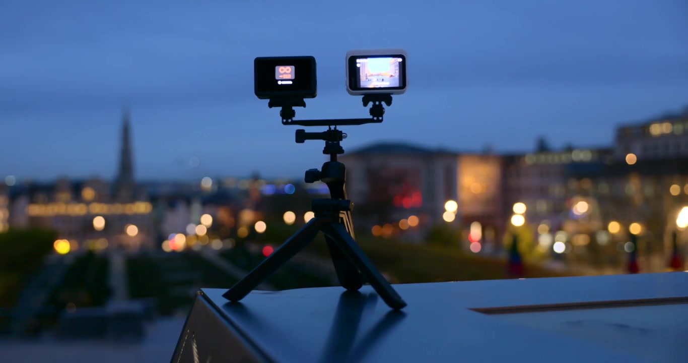 Telecamere d'azione DJI e Insta360 montate insieme su un piccolo treppiede che catturano un paesaggio urbano di notte. Primo piano, messa a fuoco selettiva.