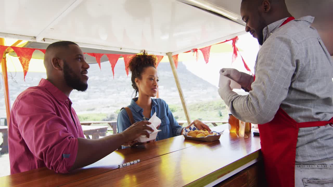아프리카계 미국인 남자가 음식 트럭에서 부부에게 햄버거와 감자를 제공하는 아프론을 입고 있습니다.