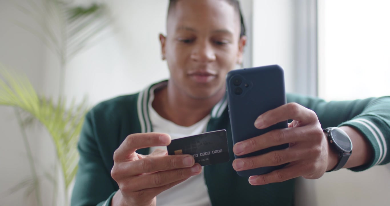 African American man in cardigan on sofa aligning card with phone scanning chip confirming payment