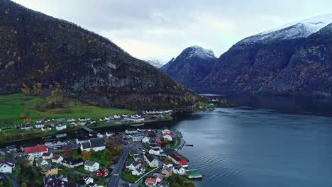 Rising-drone-flight-over-a-lakeside-village-in-a-valley-surrounded-by-snow-capped-mountains