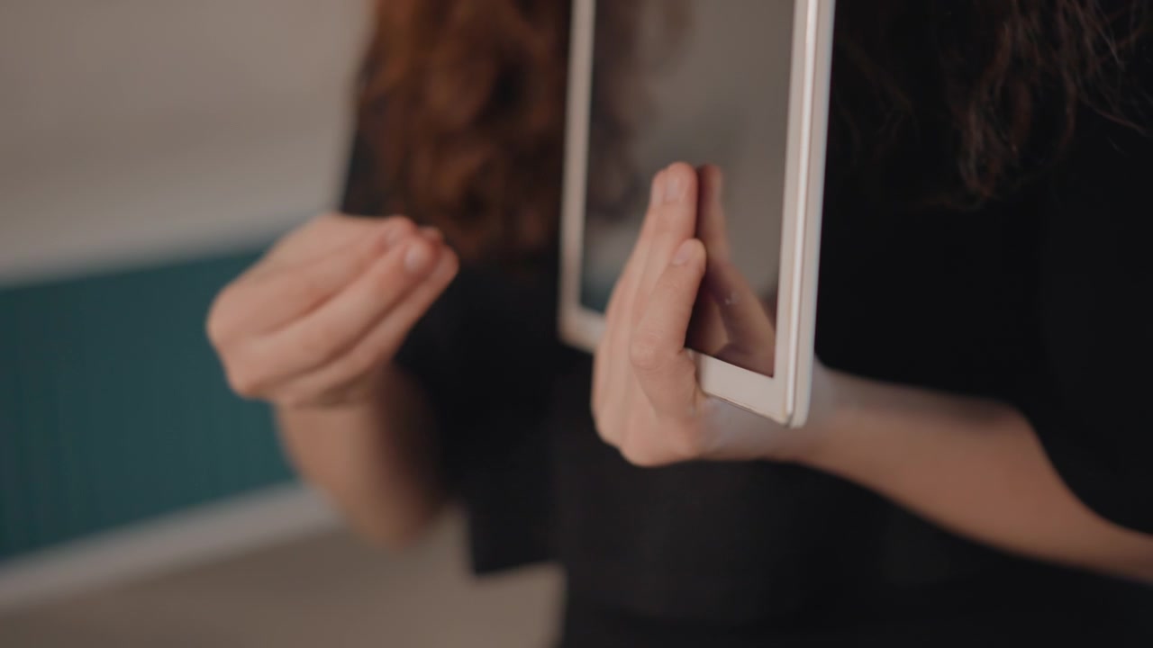 Primo piano di una persona che tiene un tablet vicino al petto, mentre aggiusta la presa e gesticola con le mani, mostrando interazione attiva, conversazione e una calma postura di presentazione al chiuso.