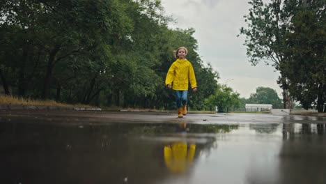 una adolescente feliz con una chaqueta amarilla corre hacia un charco y se mete en él con botas naranjas en el parque después de la lluvia