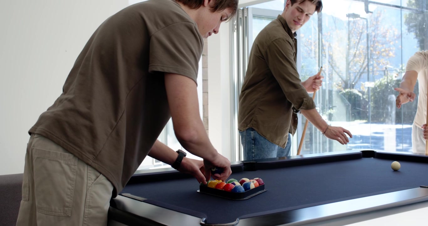 Diverse male friends positioning cue ball, urging friend racking balls at pool table starting game