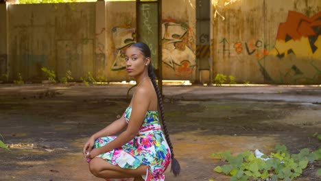 sexy young girl in a long flower dress turns while squating at an abandoned warehouse on the caribbean island of trinidad