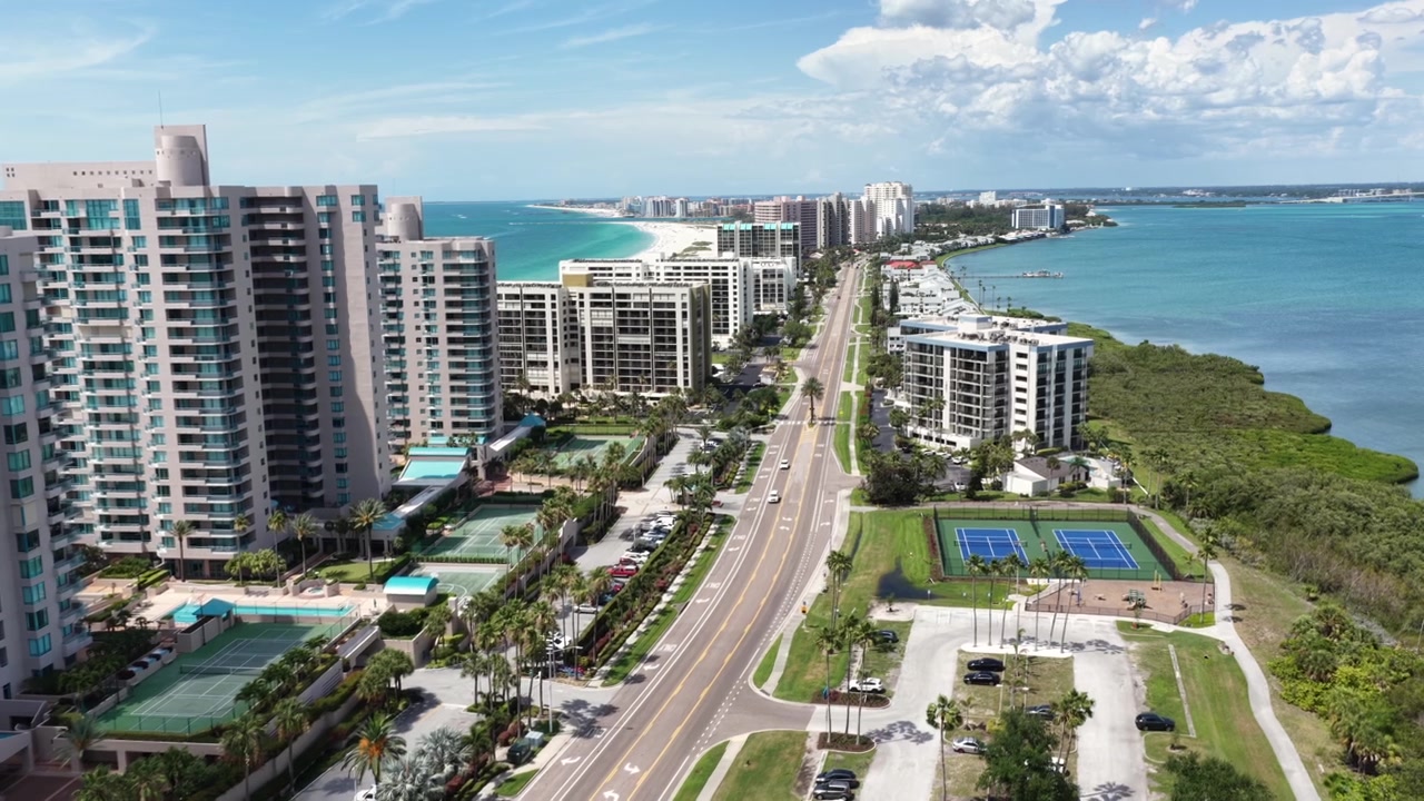 Avenida bordeada de palmeras que discurre entre torres de condominios de gran altura, canchas de tenis y estacionamientos que bordean tranquilas aguas intracosteras bajo un cielo de verano. Clearwater Beach, Florida, EE. UU. Toma aérea amplia con dron ascendente