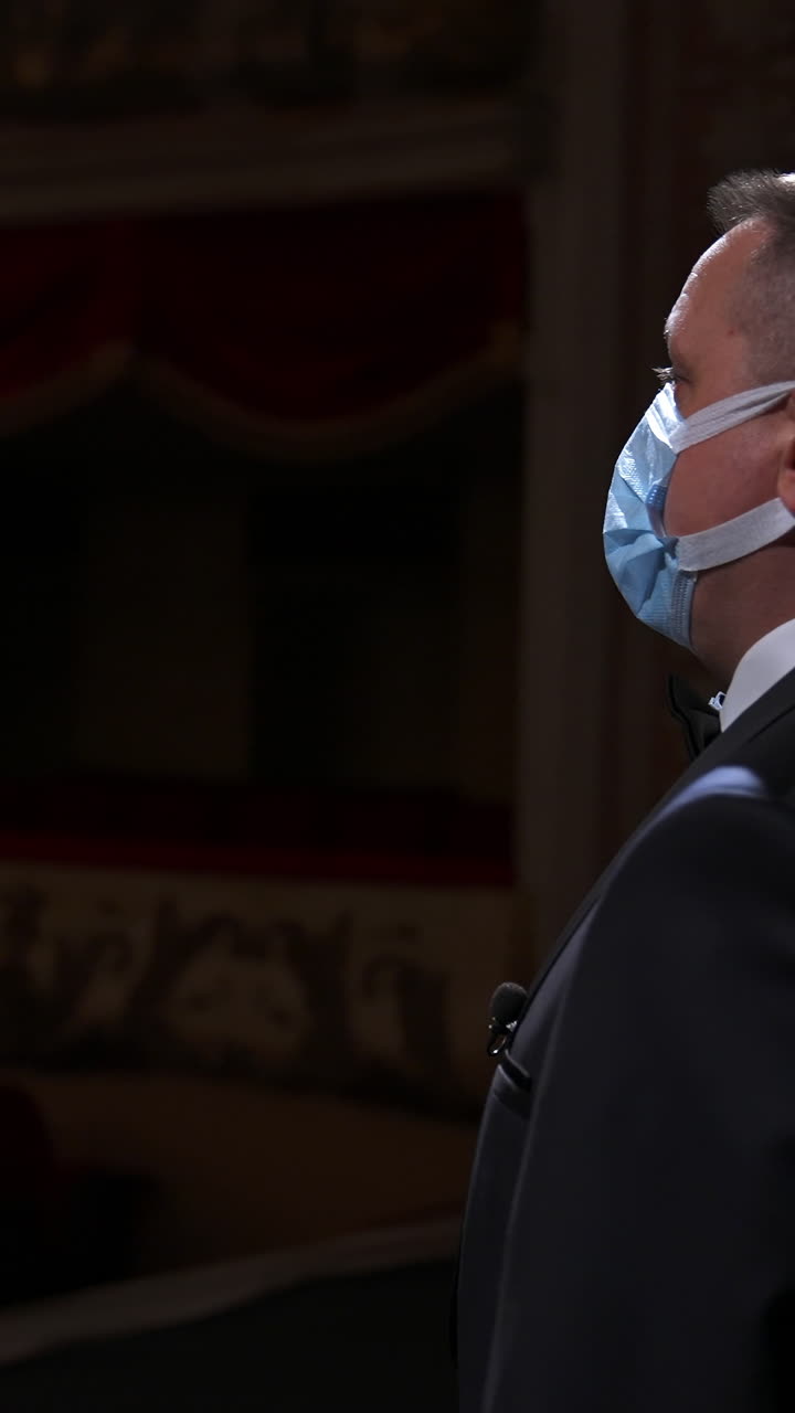 Theater actor speaks on a stage in medical mask. Announcer in suit stands in front of empty theater hall. Pandemic concept. Vertical video
