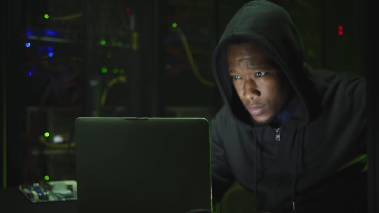 African american male computer hacker using laptop in business server room