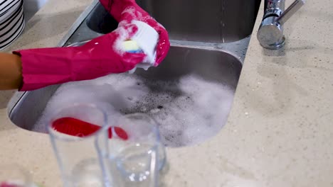 hands in pink gloves wash a bowl in a kitchen sink filled with soapy water. bright lighting enhances the clean, modern setting