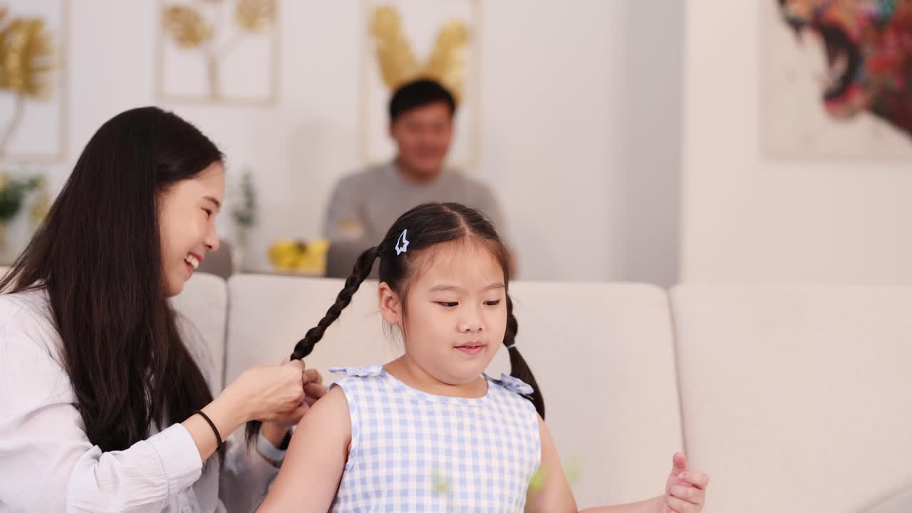 A mother braids her daughter's hair in a bright, cozy living room with a relaxed atmosphere