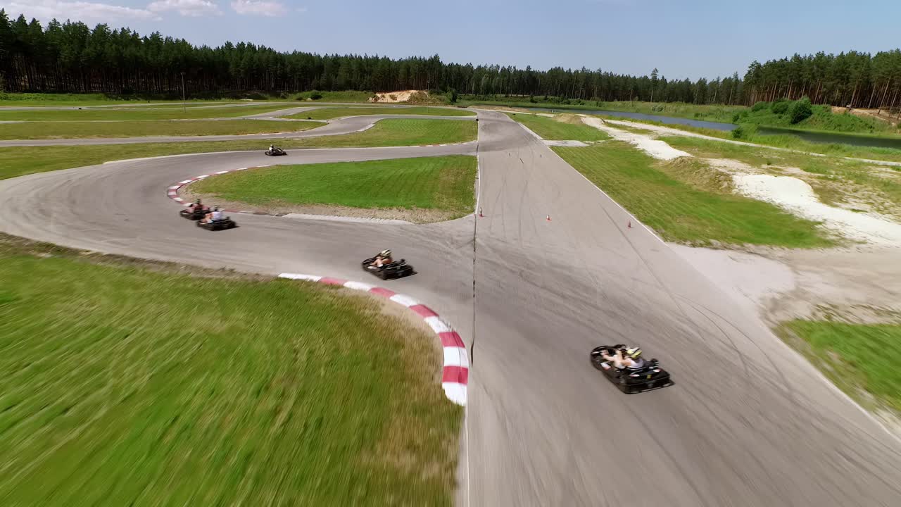 Sporta Komplekss 333, sunny summer day, aerial shot with wide-angle lens, capturing go-karts racing on the gray asphalt track, surrounded by vibrant green fields and a dense forest backdrop