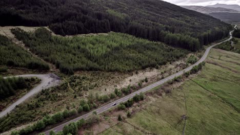 Single-Car-Drives-Down-Winding-Road-Along-Dark-Moody-Woods,-Scottish-Highlands