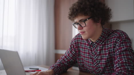 un hombre rizado con una mirada seria trabaja en una computadora portátil sentado en una cocina moderna. joven estudiante independiente usando computadora portátil estudiando en línea trabajando desde casa en internet