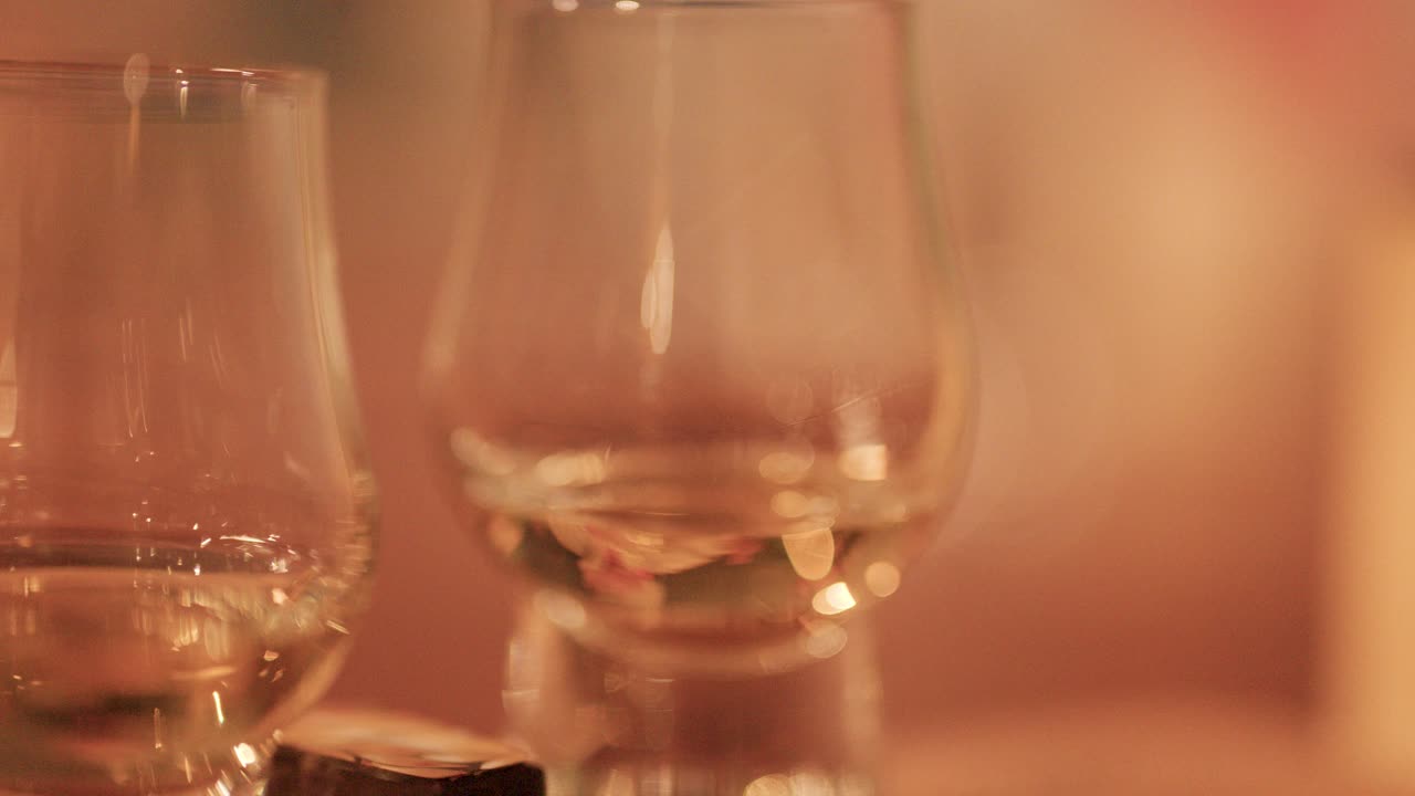 En hånd løfter et whiskyglas fra et bord ved siden af chokolade, med blød, varm omgivende belysning og lav dybdeskarphed. Kameraet forbliver stabilt.