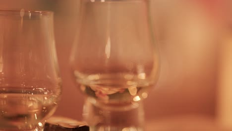 a hand lifts a whisky glass from a table beside chocolate, with soft, warm ambient lighting and a shallow depth of field. camera remains steady