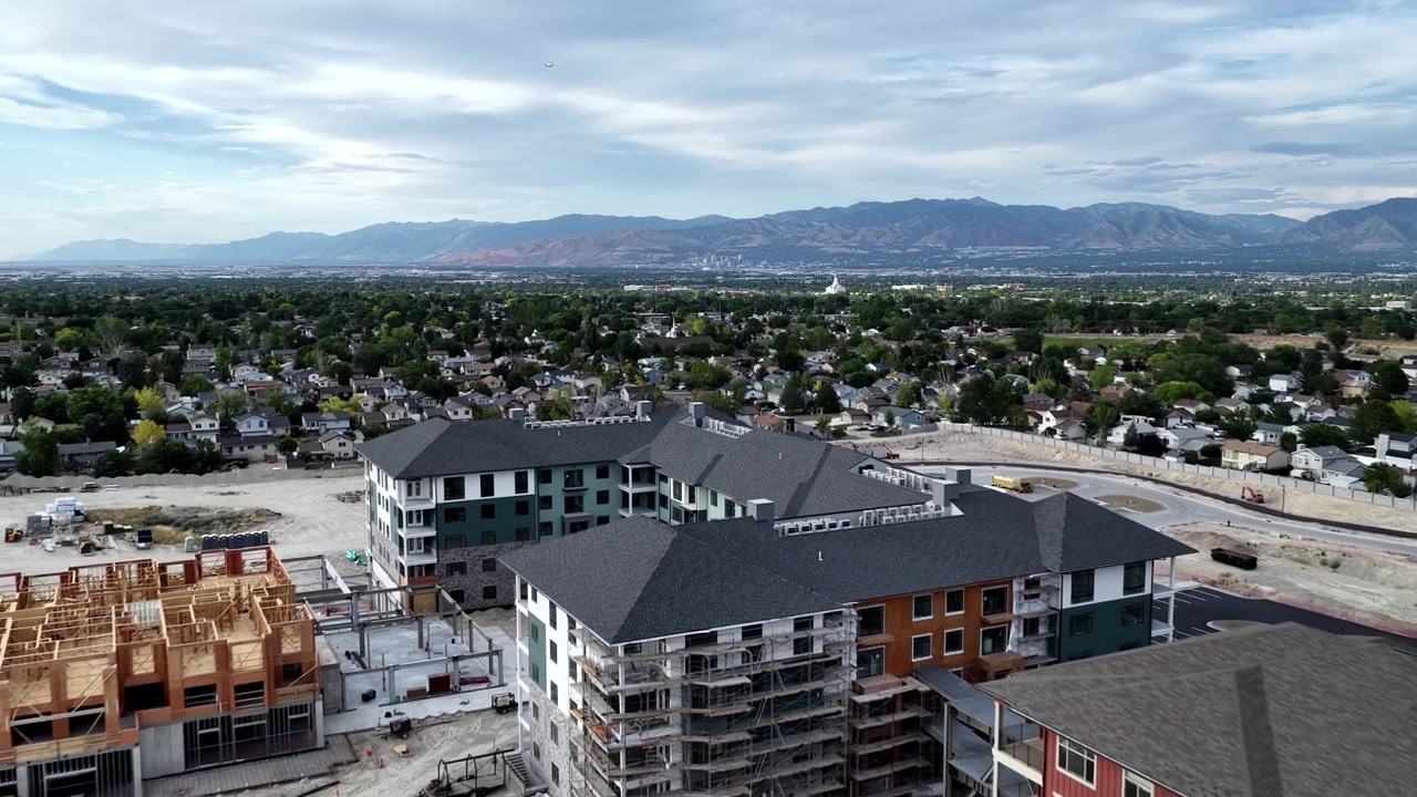 Apartment construction project beside completed multifamily buildings in Taylorsville, Salt Lake County, Utah, USA, beneath Wasatch Range with active earthworks and tower crane, drone forward flyover