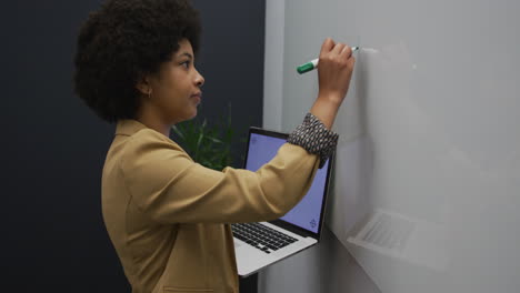 biracial businesswoman standing using laptop writing on a board in modern office