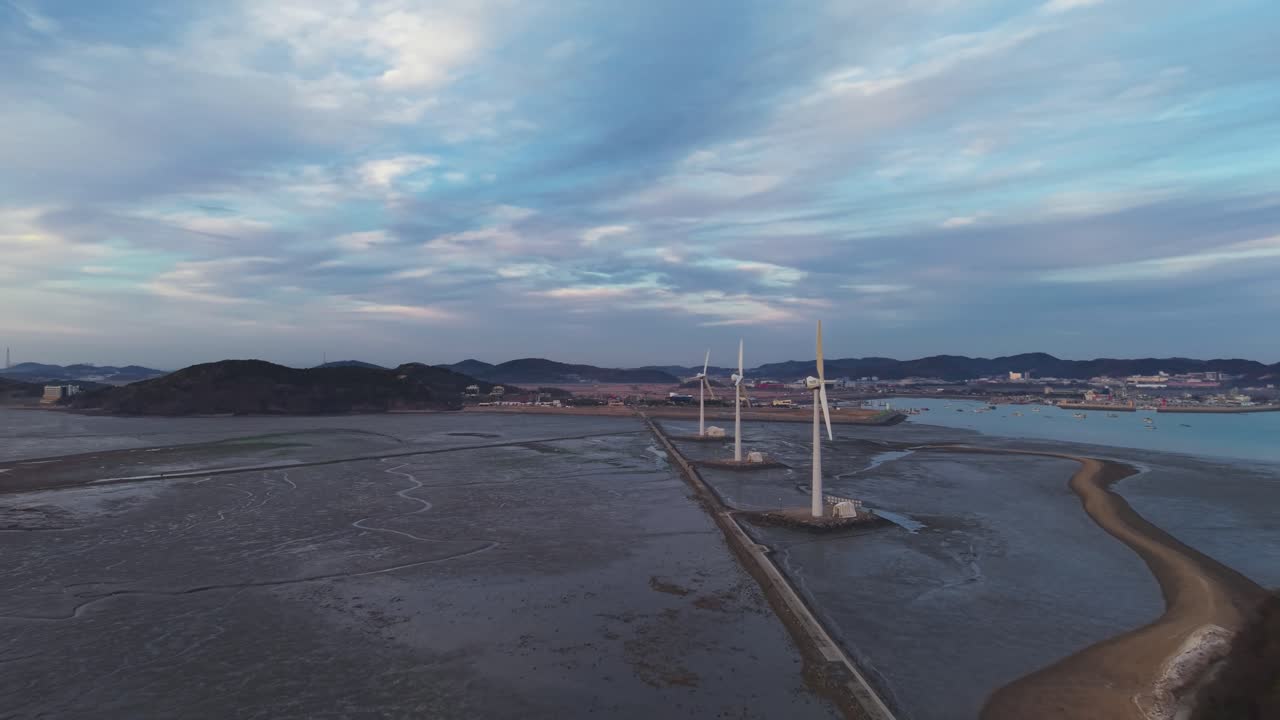 대한민국 안산의 흐린 하늘 아래, 썰물 때 갯벌 위에 세 개의 풍력 터빈이 서 있고 누에섬으로 이어지는 탄도 바닷길을 광활하게 항공 촬영한 모습.