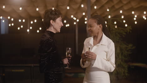 two elegant multiethnic women holding champagne glasses and talking to each other at new year's eve party