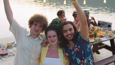 portrait of joyous friends embracing and waving at lake party