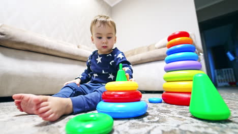 calm pretty baby boy sitting on the floor with his toys around. cute toddler disassembles the pyramid. low angle view.