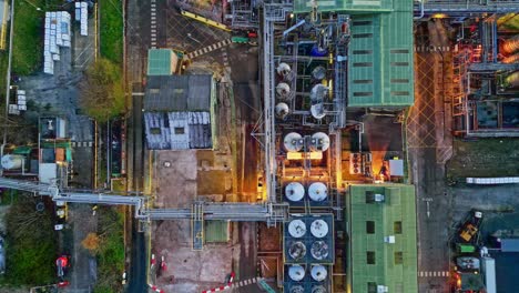 slow sideways moving aerial drone footage of a large industrial plant at dusk, showing pipework structures, buildings, cooling towers, steam, and work vehicles