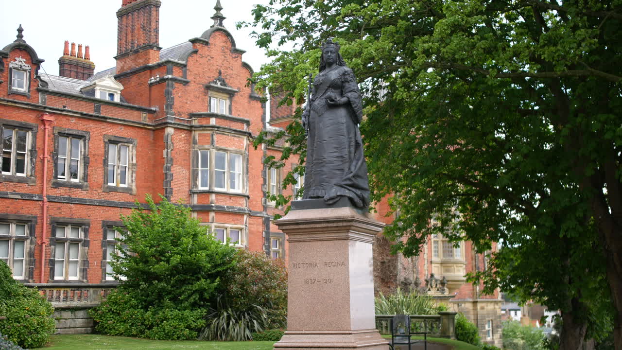 A bronze statue of Queen Victoria standing on a stone pedestal in front of a red-brick historic building and leafy trees in Scarborough, North Yorkshire in England