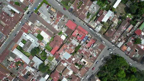 Drone-aerial-footage-of-car-and-motorbike-street-traffic-in-Antigua,-Guatemala-colonial-town-showing-bright-colorful-red-rooftops,-and-green-tree-tops