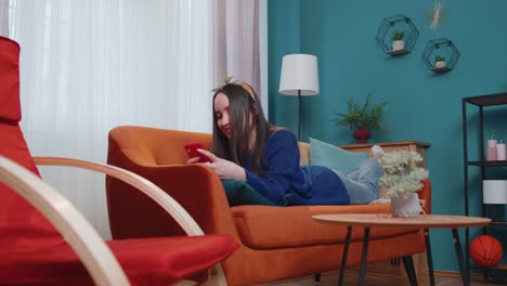mujer joven caucásica relajada y sonriente acostada en el sofá de su casa con un teléfono inteligente desplazándose, navegando en línea