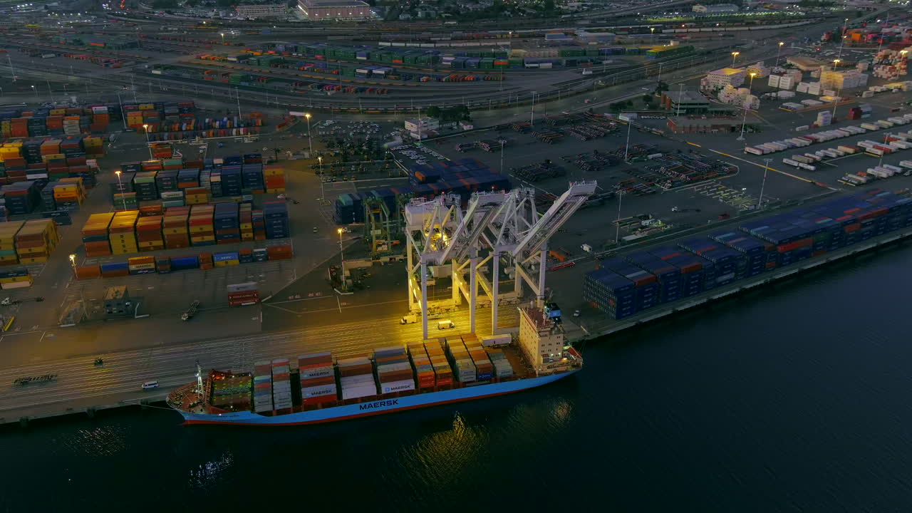 The glowing lights from a crane and ship at the Port of Oakland at nighttime - aerial parallax tilt down view