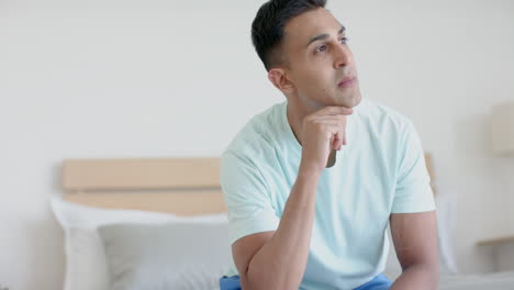 Contemplative-biracial-man-sitting-on-bed-in-sunny-bedroom-thinking,-slow-motion