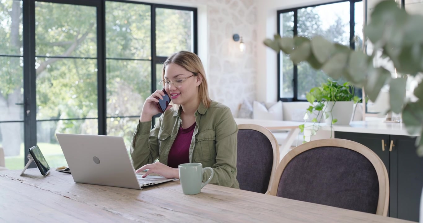 Donna che risponde a una chiamata sullo smartphone mentre digita sul laptop al tavolo da pranzo, lavorando da remoto