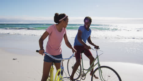 african american couple smiling and riding bikes on the beach