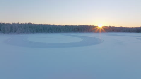 Curving-tyre-tracks-on-Norbotten-Polar-circle-woodland-ice-lake-aerial-view-rising-to-glowing-sunrise-skyline