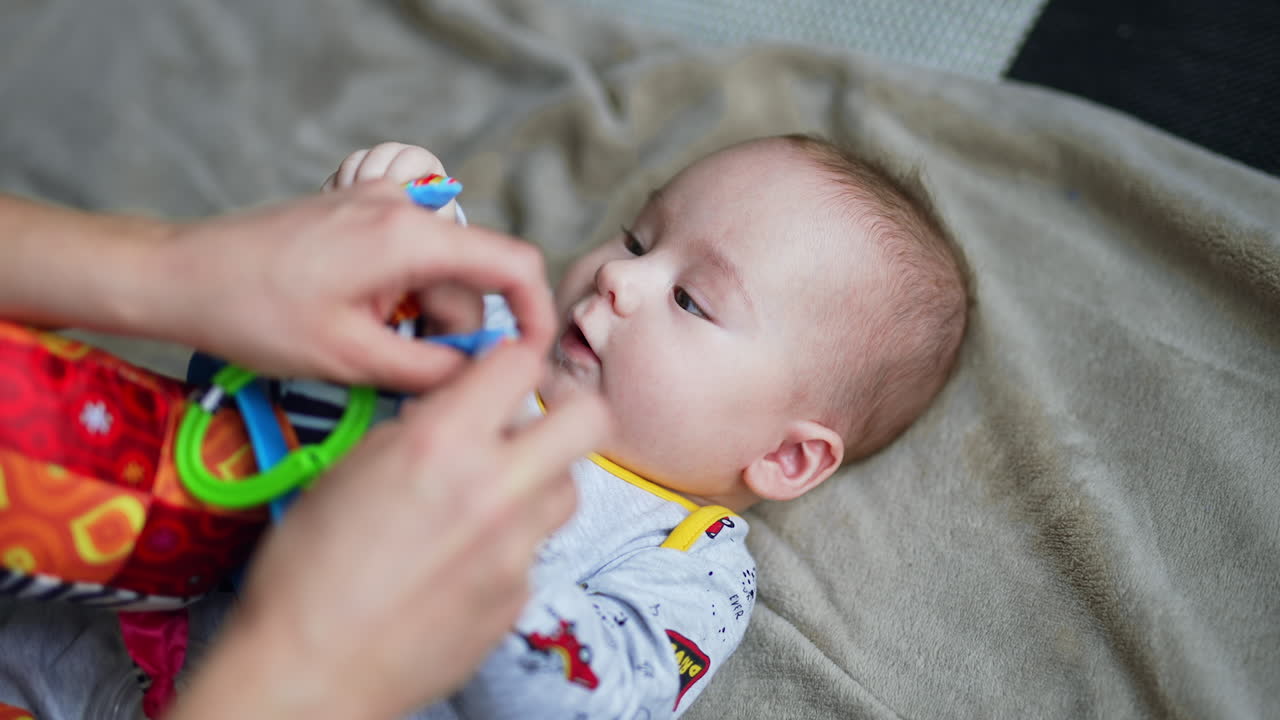 Mother is giving a bright soft toy to a baby boy lying on his back. Cute little child pulls the toy into mouth. Close up.