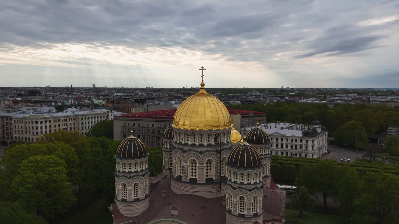 Riga’s Nativity Cathedral stands tall in a central park, backed by cityscape and sky.
