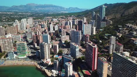 Luftaufnahme-Von-Wohngebäuden-In-Der-Nähe-Der-Küste,-Umgeben-Von-Strand-Und-Ozeanwasser-Mit-Bergen-An-Einem-Klaren-Himmelstag-In-Der-Stadt-Benidorm-In-Spanien