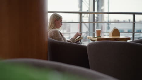 side view of woman seated, reading a book thoughtfully turns to a new page with coffee cup on the table, modern cafe with blurred outdoor urban view