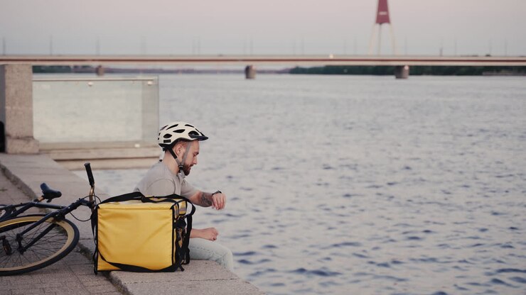 Food Delivery Man Rests Next To His Thermal Backpack At Sunset On The Shore Of A River And Consults At His Smartwatch