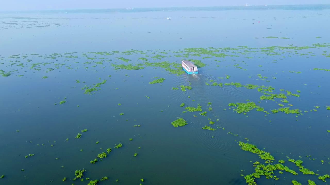 Passenger ferry boat cruising through vast backwaters covered with patches of water hyacinth and aquatic plants, wide aerial shot