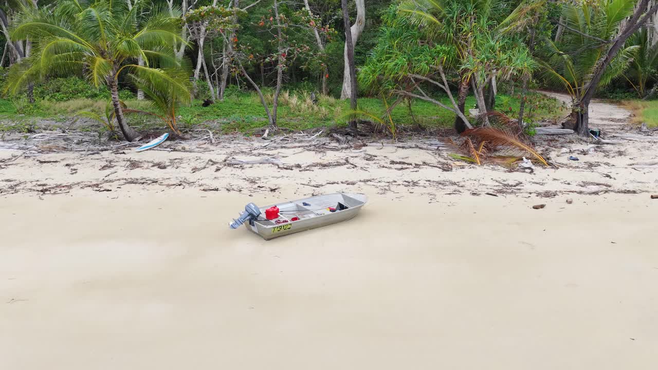 Drone flies toward small abandoned motorboat on sandy beach, lush rainforest background, overcast daylight