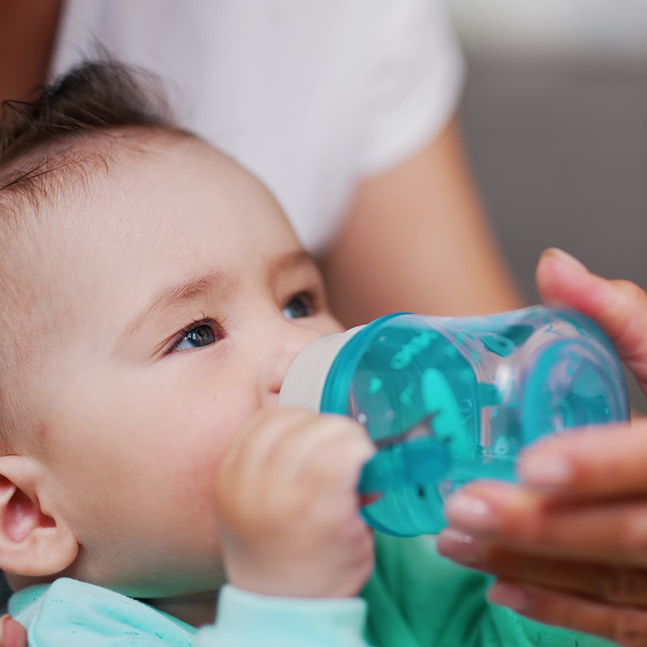 Adorable baby sits in mom's arms holding a bottle. Sweet Caucasian child drinking water. Close up