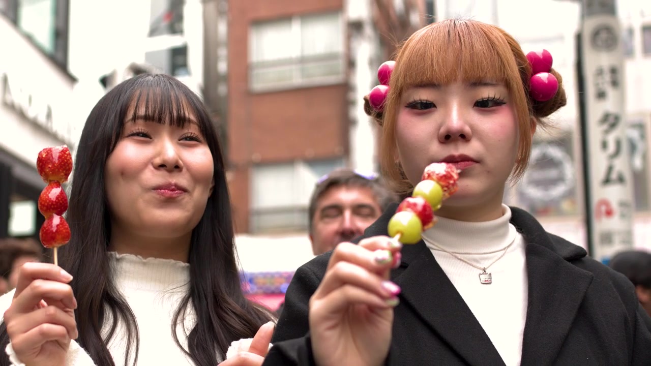 Due donne giapponesi alla moda gustano spiedini di fragole su una strada affollata di Tokyo durante il giorno.