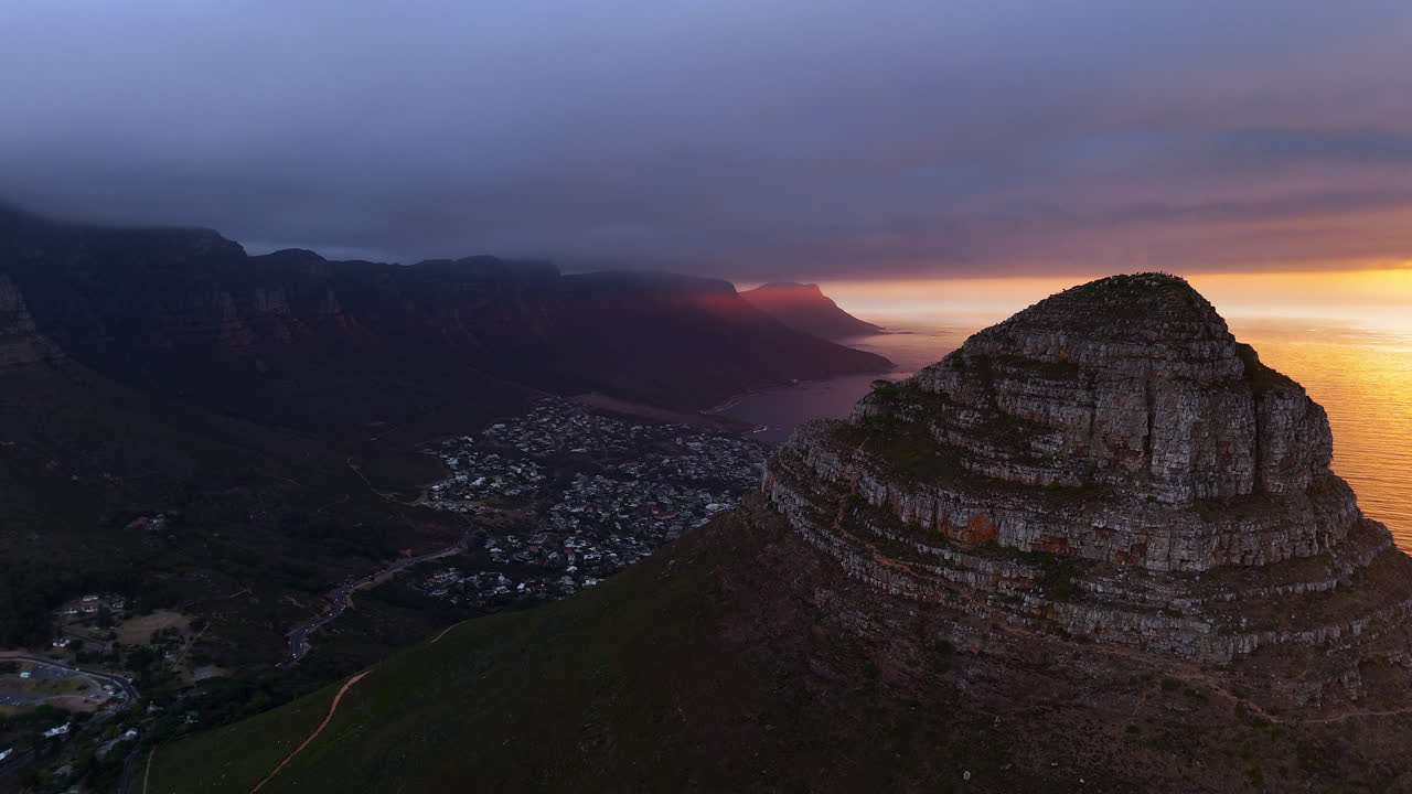 Cinematic aerial view of Table Mountain in Cape Town, South Africa, dramatic rocky cliffs with coastal cityscape at sunset, clouds rolling over peaks and golden evening light