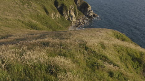 Epic-view-of-a-seaside-coast-line-with-big-steep-cliffs,-sea-waves-washing-ashore-and-beautiful-green-inclined-fields-with-high-grass,-and-reveal-of-a-sea-port-in-the-distance,-on-the-sunset