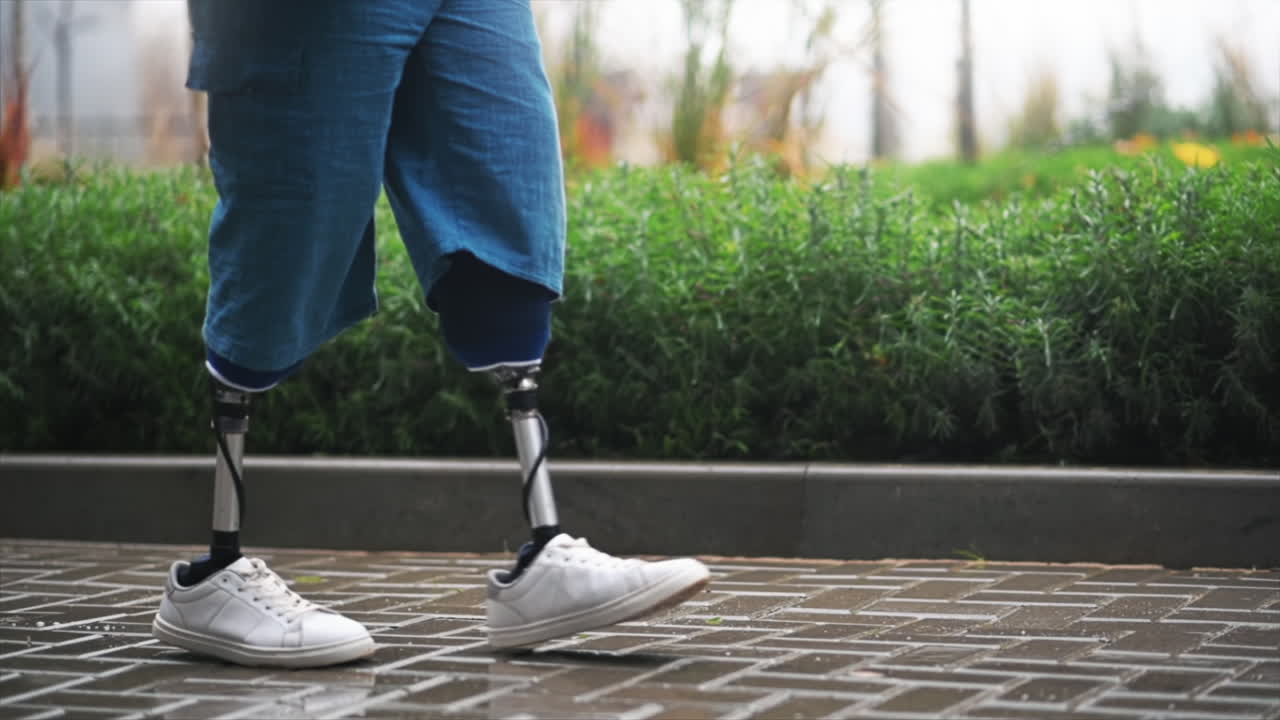 View of a walking man with prosthetic legs and white sneakers at rainy weather, greenery on the background